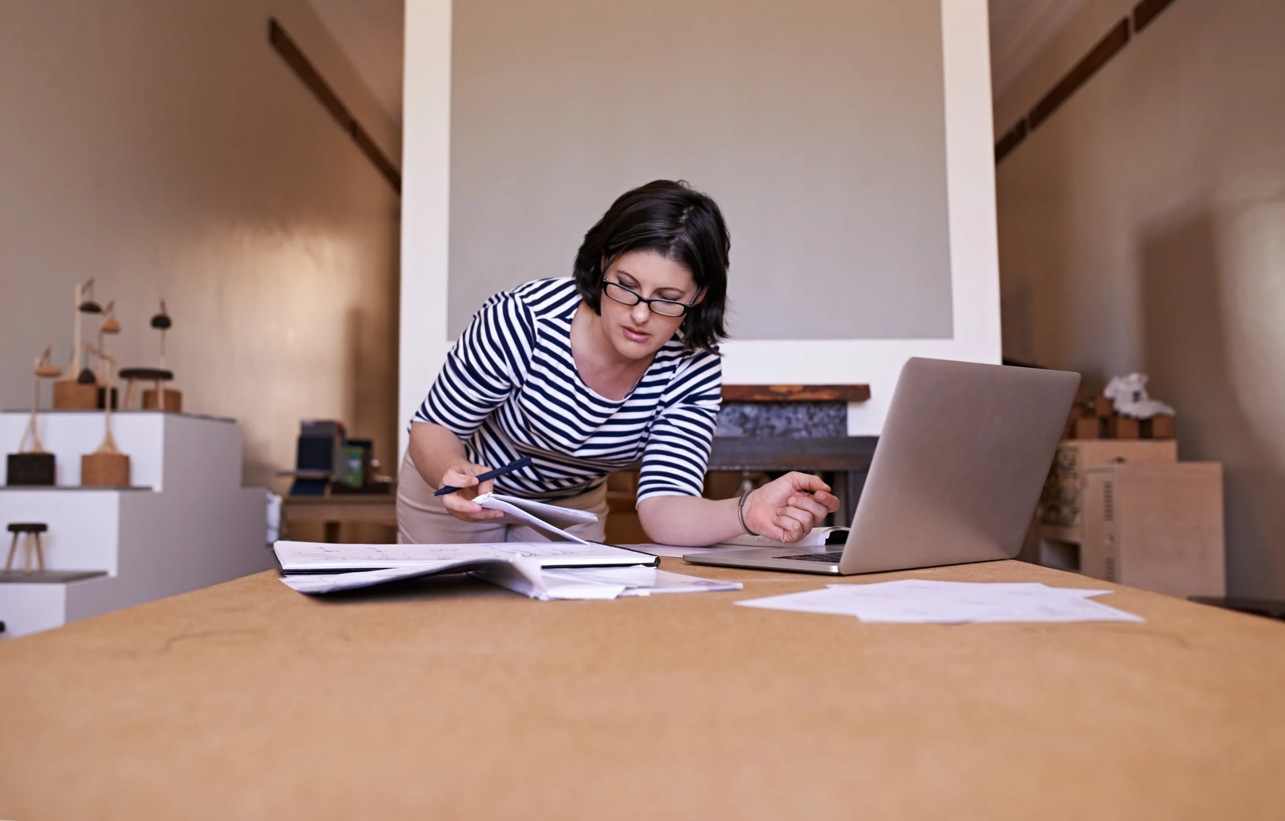 Woman with dark shoulder length hair, wearing a white and black t-shirt and black rimmed glasses is leaning over her desk with paperwork and laptop preparing overdue tax returns