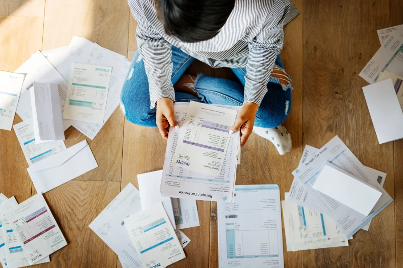 Woman sitting on a wooden floor sorting invoices scattered around her.