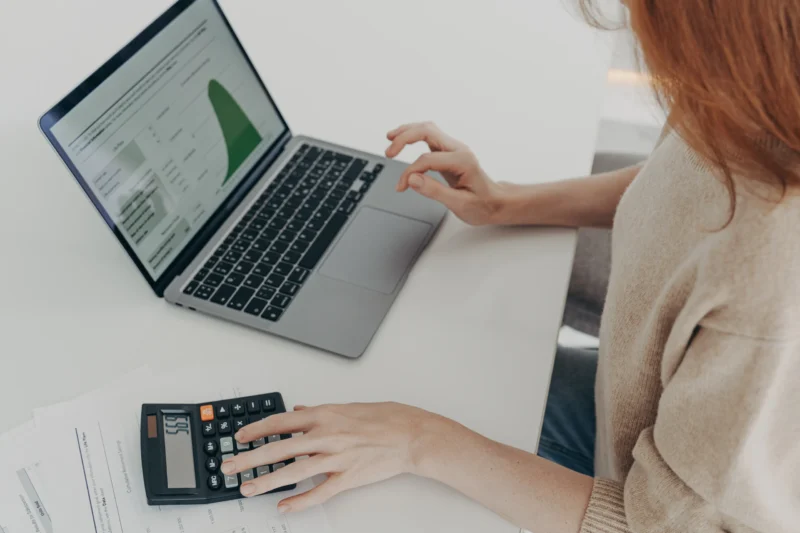 Woman using laptop to look at results from a tax return calculator. She also has an old school calculator next to her.