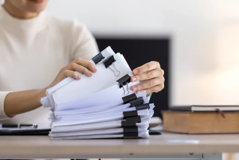Woman sorting a large stack of paperwork at a desk with a computer.